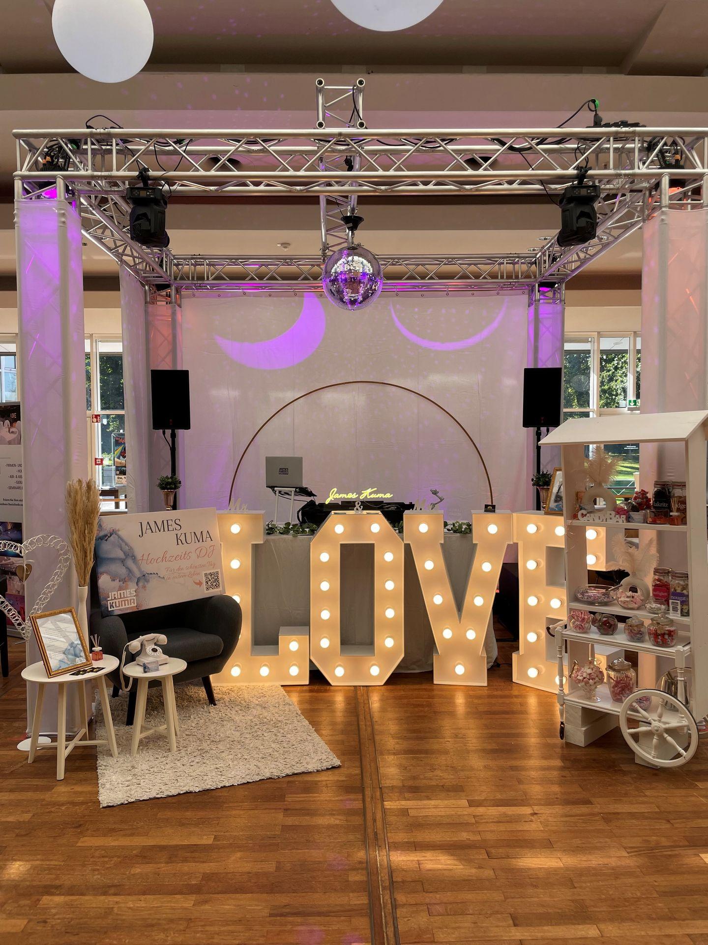 Wedding DJ setup with large illuminated LOVE sign and disco ball in decorated event space.