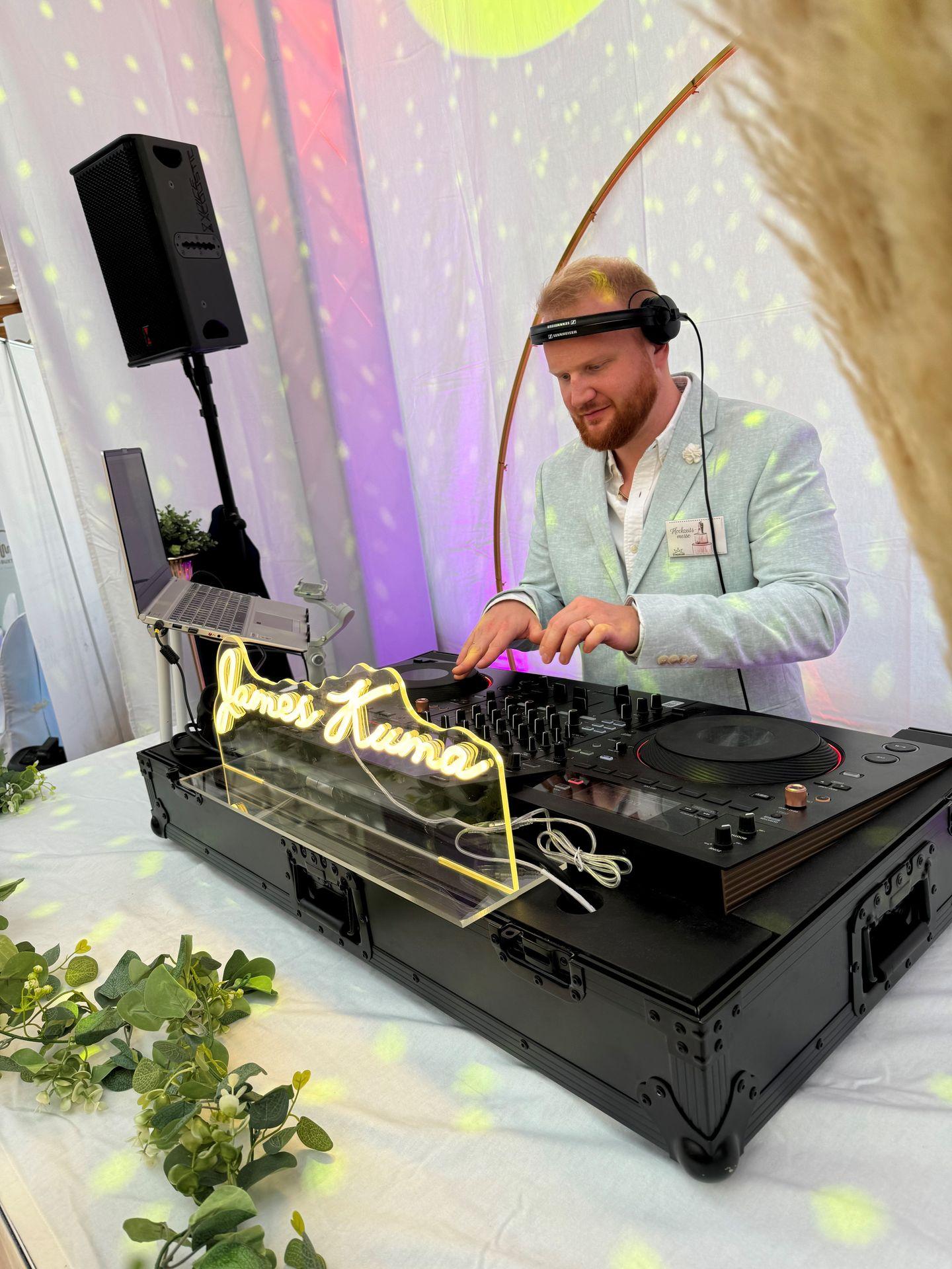 DJ performing at a setup with a neon sign, surrounded by greenery and illuminated by colorful lights.