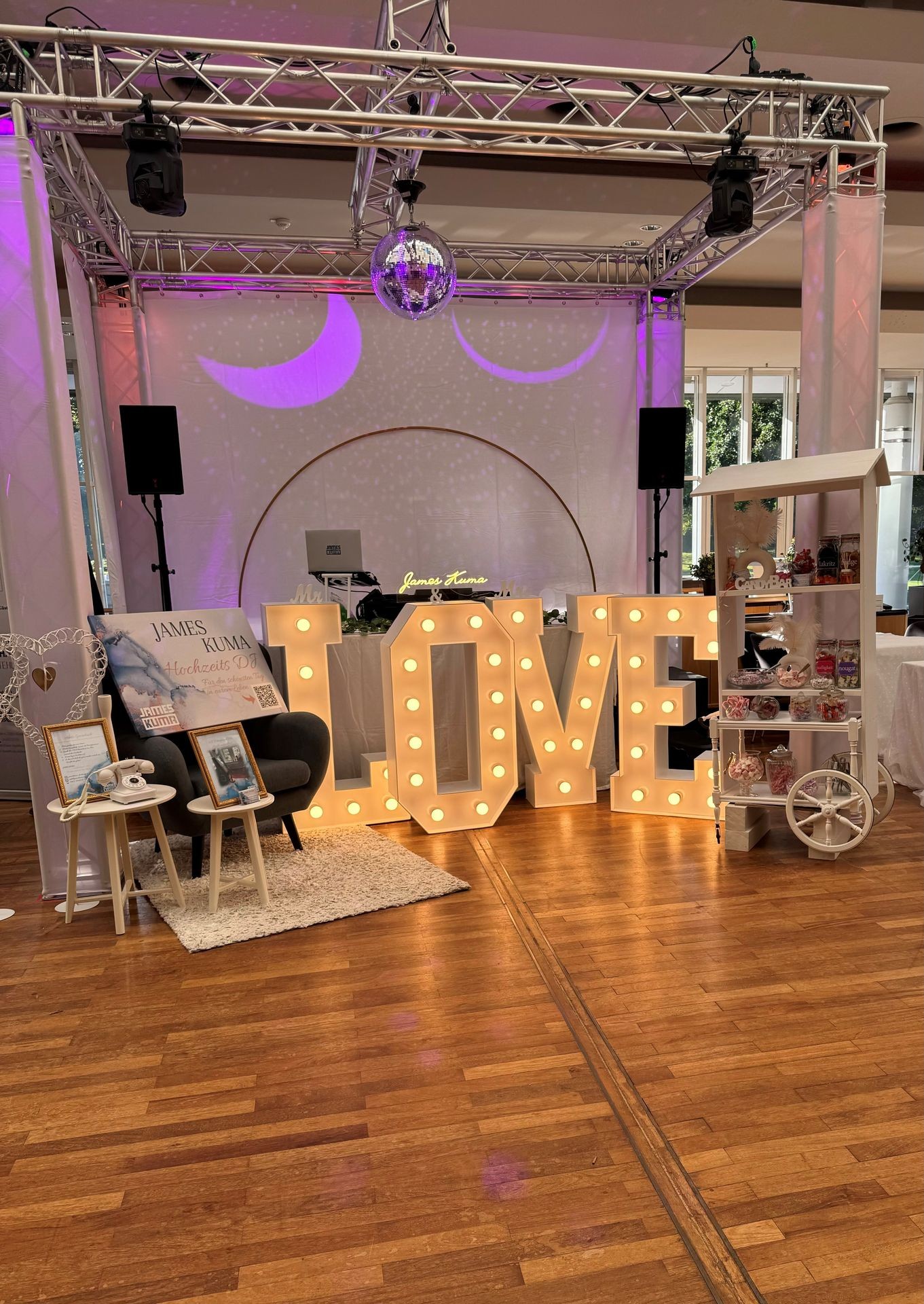 Wedding decoration with large illuminated LOVE letters and candy cart on wooden floor.