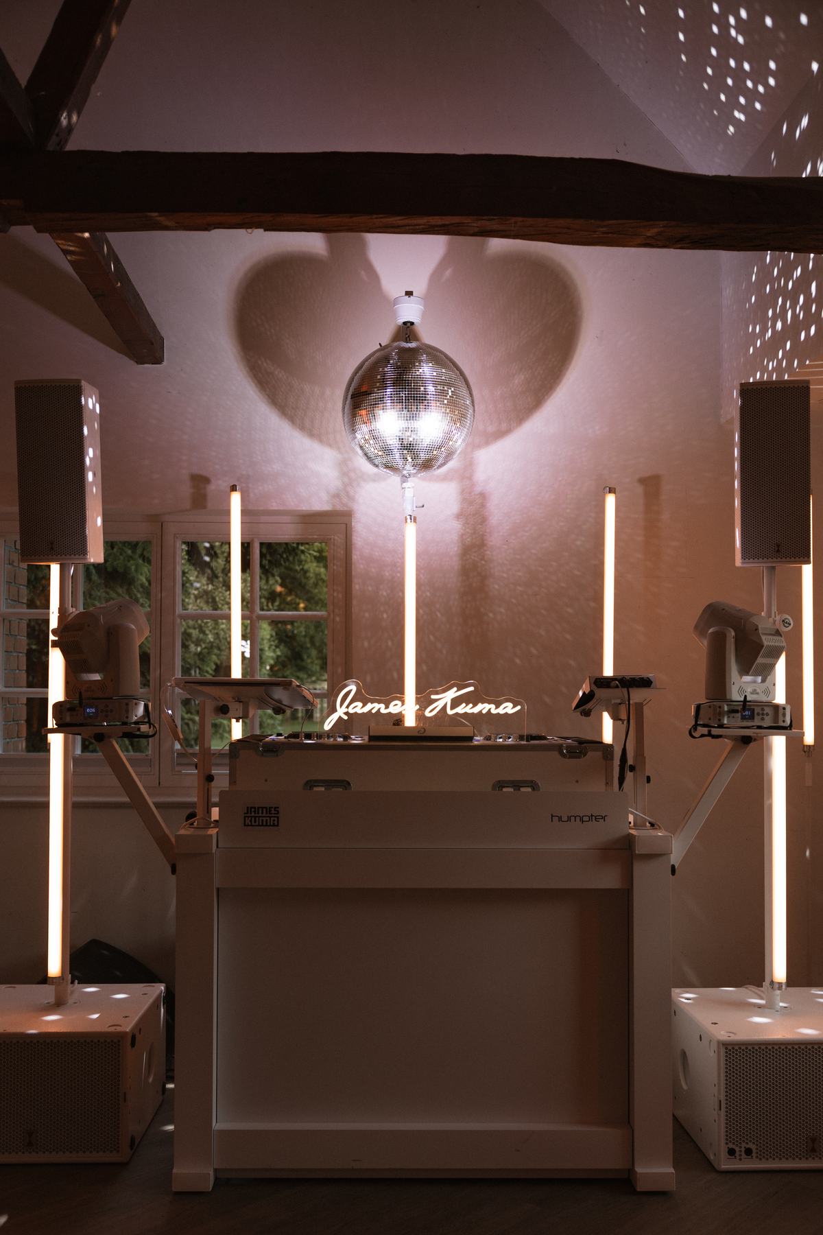 Weiße DJ-Booth mit leuchtendem Namenszug, Discokugel mit herzförmigem Schatten an der Wand