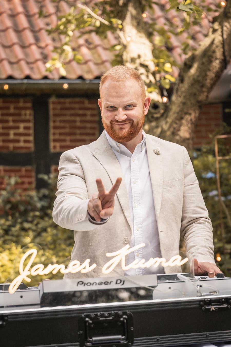 James Kuma im hellen Anzug hinter den Decks unter freiem Himmel, vor ihm der leuchtende Namenszug