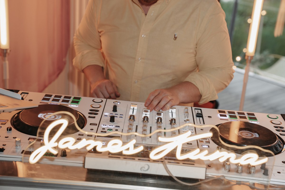 Hände am Mixer hinter dem leuchtenden Neon-Schriftzug James Kuma auf den weißen Decks