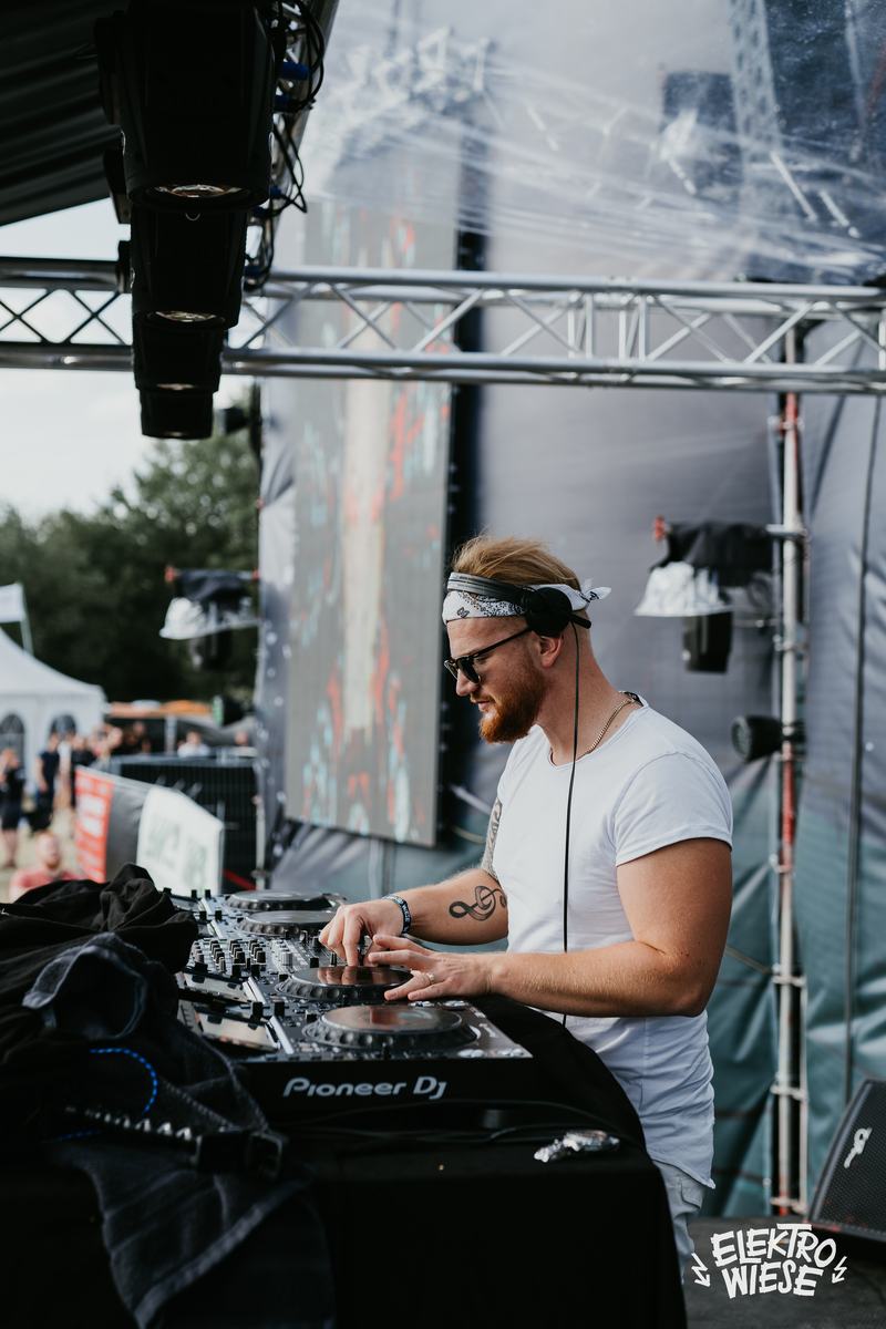 James Kuma mit Kopfhörern und Sonnenbrille an den Decks einer Festivalbühne, Traversen im Hintergrund