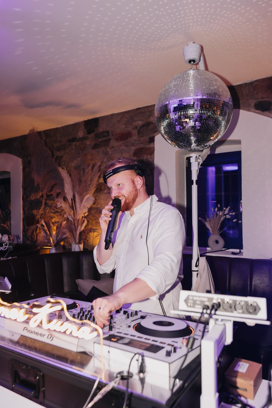James Kuma am Mikrofon hinter den weißen Decks, über ihm die Discokugel vor einer Natursteinwand