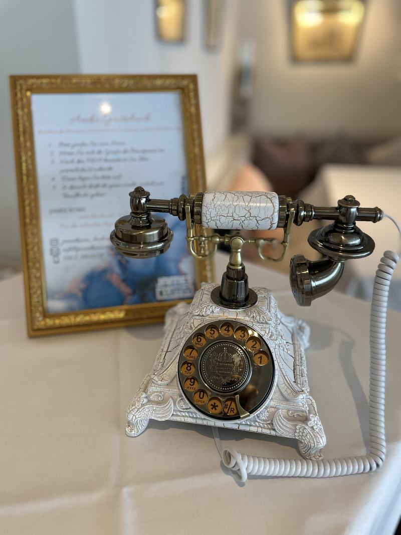 Weißes Retro-Telefon mit Wählscheibe als Audio-Gästebuch, daneben die gerahmte Anleitung für die Gäste